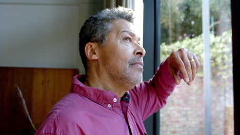 Biracial-senior-man-leaning-on-window-and-looking-through-it-at-home