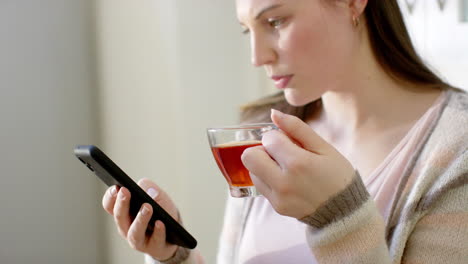 Mujer-Caucásica-Usando-Un-Teléfono-Inteligente-Y-Bebiendo-Té-En-Casa,-En-Cámara-Lenta
