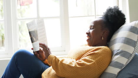 Happy-african-american-senior-woman-lying,-reading-book-and-smiling-in-sunny-room,-slow-motion