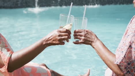 Midsection-of-senior-african-american-female-friends-sitting-by-pool-making-a-toast,-slow-motion
