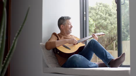 Happy-senior-biracial-man-sitting-by-open-window-playing-guitar-at-home,-copy-space,-slow-motion