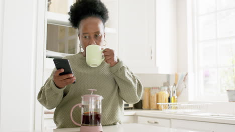 Feliz-Mujer-Mayor-Afroamericana-Tomando-Café,-Usando-Un-Teléfono-Inteligente-En-Una-Cocina-Soleada,-Cámara-Lenta