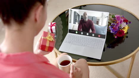 Caucasian-woman-holding-red-gift-using-laptop-with-african-american-man-on-screen