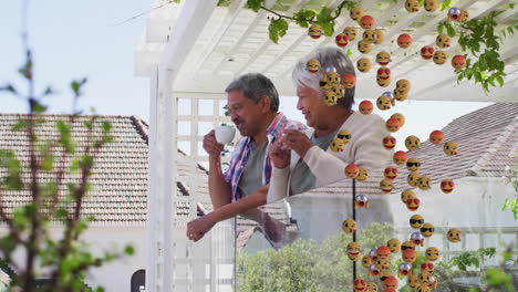 Animation-of-emoji-icons-over-senior-biracial-couple-drinking-coffee-on-balcony