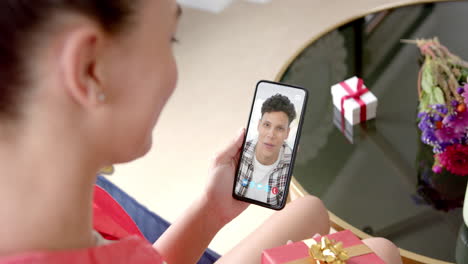Biracial-woman-holding-smartphone-with-biracial-man-on-screen-with-gift-on-desk