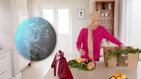 Biracial-woman-in-hijab-taking-vegetables-out-of-box-in-kitchen-over-globe