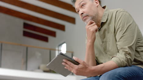 Thoughtful-senior-biracial-man-sitting-on-table-using-tablet-at-home,-copy-space,-slow-motion