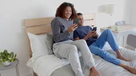 Happy-diverse-gay-male-couple-sitting-on-bed-using-smartphones-in-the-morning,-slow-motion