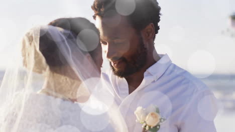 Animation-of-light-spots-over-happy-african-american-groom-unveiling-bride-on-beach-at-wedding