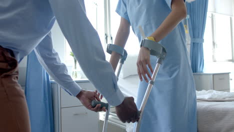 Midsection-of-diverse-male-doctor-giving-female-patient-crutches-in-hospital-ward,-slow-motion