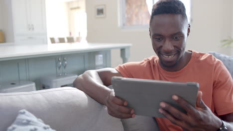 Glücklicher-Afroamerikanischer-Mann,-Der-Im-Sonnigen-Wohnzimmer-Sitzt-Und-Ein-Tablet-Benutzt,-Zeitlupe