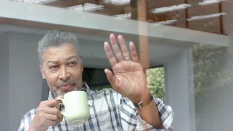 Happy-senior-biracial-man-waving-through-window-and-drinking-coffee-at-home,-copy-space,-slow-motion