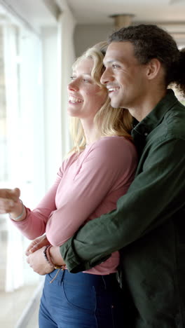 Vertical-video-of-happy-diverse-couple-embracing-at-window-at-home,-slow-motion