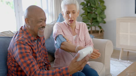 Happy-senior-biracial-couple-sitting-on-couch-and-taking-pressure-at-home,-slow-motion