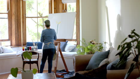 Senior-biracial-woman-wearing-apron-and-painting-on-big-canvas-at-home,-slow-motion
