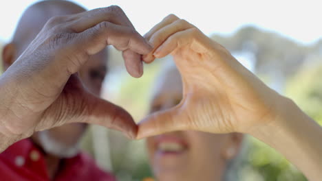 Happy-senior-biracial-couple-making-heart-gesture-in-sunny-garden-at-home,-slow-motion