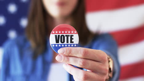 Caucasian-woman-holds-a-"VOTE"-badge,-American-flag-in-the-background