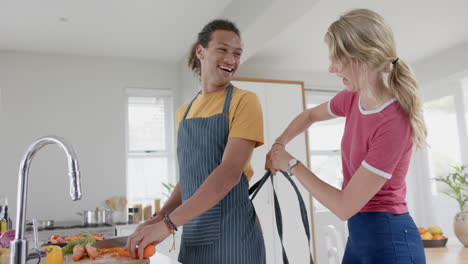 Happy-diverse-couple-preparing-fresh-vegetables-and-wearing-aprons-in-kitchen,-slow-motion