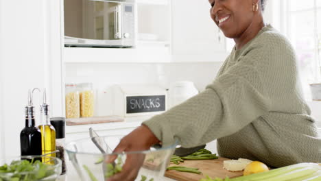 Glückliche-Afroamerikanische-Seniorin,-Die-Gemüse-Hackt-Und-In-Der-Sonnigen-Küche-Lächelt,-Zeitlupe