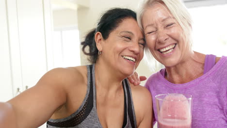 Two-happy-diverse-senior-women-with-cocktail-doing-selfie-laughing-in-kitchen,-slow-motion