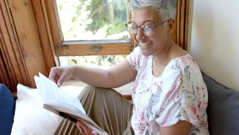 Happy-senior-biracial-woman-on-couch-reading-book-at-window-at-home,-slow-motion