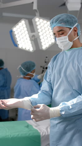 Vertical-video-of-biracial-male-surgeon-wearing-medical-gloves-in-operating-theatre,-slow-motion