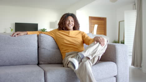 Portrait-of-happy-biracial-man-with-long-hair-sitting-on-sofa-smiling-in-living-room,-slow-motion