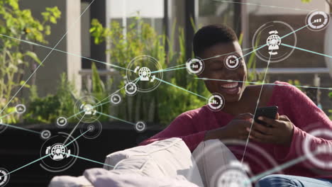 Animation-of-network-of-connections-with-icons-over-african-american-woman-using-smartphone