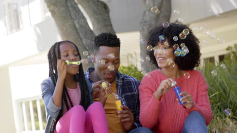 Glückliche-Afroamerikanische-Eltern-Mit-Tochter,-Die-Zu-Hause-Im-Garten-Seifenblasen-Pusten,-Zeitlupe