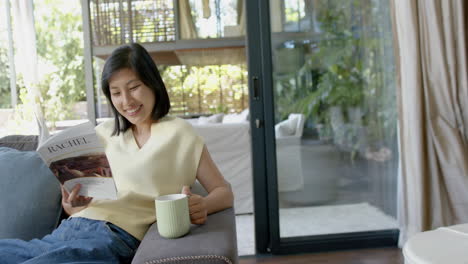 Glückliche-Asiatische-Frau,-Die-Mit-Einer-Tasse-Auf-Der-Couch-Sitzt-Und-Ein-Buch-Im-Sonnigen-Wohnzimmer-Liest,-Zeitlupe