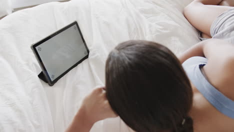 Biracial-woman-lying-on-bed-using-tablet-with-copy-space-on-screen,-slow-motion