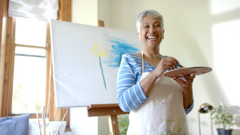 Portrait-of-happy-senior-biracial-woman-mixing-colours-on-palette-at-home,-slow-motion