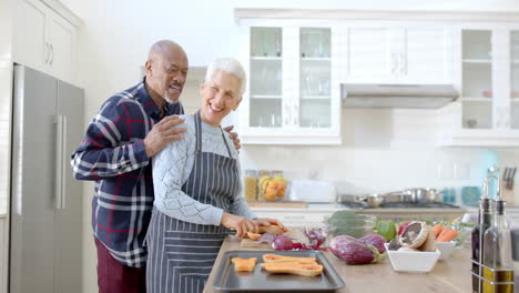 Happy-senior-diverse-couple-preparing-vegetables-and-embracing-in-kitchen,-copy-space,-slow-motion