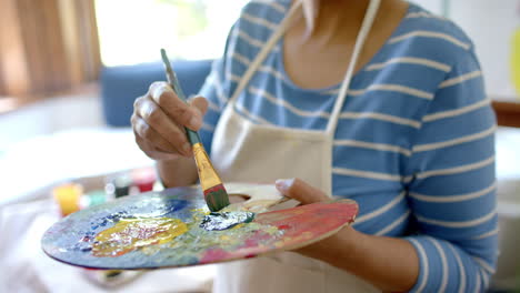 Portrait-of-happy-senior-biracial-woman-mixing-colours-on-palette-at-home,-slow-motion