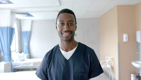 Portrait-of-happy-african-american-male-doctor-in-scrubs-smiling-in-hospital-ward,-slow-motion