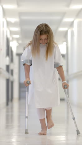 Vertical-video-of-caucasian-girl-patient-walking-with-crutches-in-hospital-corridor,-slow-motion