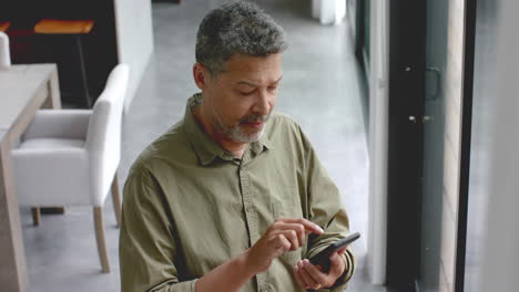 Happy-senior-biracial-man-using-smartphone-standing-by-window-at-home,-slow-motion