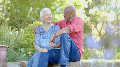 Happy-senior-biracial-couple-sitting-on-wall-and-talking-in-sunny-garden-at-home,-slow-motion