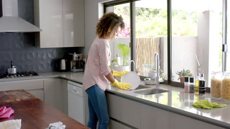 Happy-biracial-woman-washing-up-dishes-at-sink-in-sunny-kitchen,-copy-space,-slow-motion