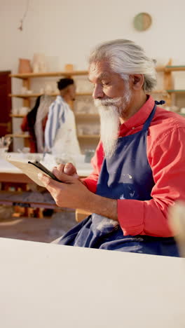 Alfarero-Birracial-Enfocado-Con-Barba-Larga-Usando-Tableta-En-Estudio-De-Cerámica,-Cámara-Lenta