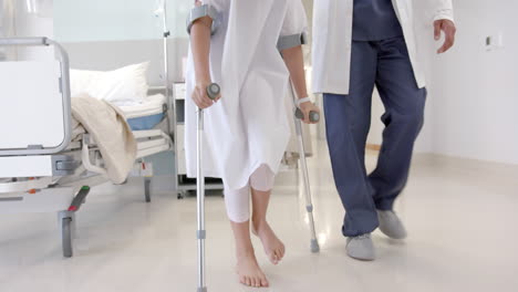 Low-section-of-diverse-male-doctor-helping-girl-patient-use-crutches-in-hospital-ward,-slow-motion