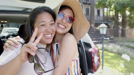 Happy-asian-female-friends-with-sunglasses-and-sun-hat-sitting-in-car-trunk-and-smiling,-slow-motion