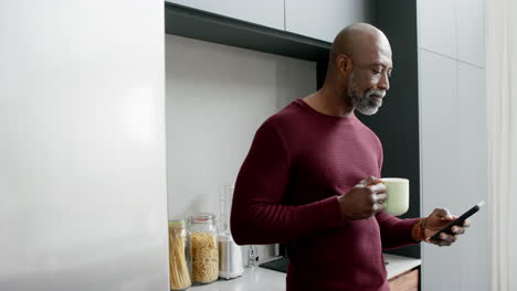 Happy-biracial-man-drinking-coffee-and-using-smartphone-in-kitchen,-slow-motion