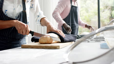 Biracial-couple-wearing-aprons-and-cutting-fresh-bread-in-kitchen,-slow-motion