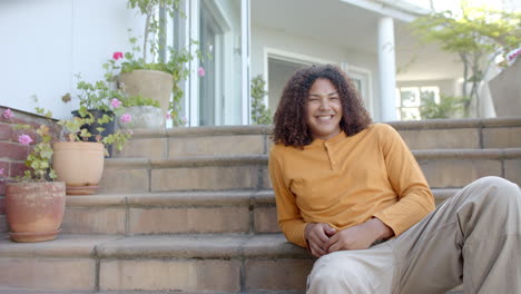 Portrait-of-happy-biracial-man-with-long-hair-sitting-on-steps-in-garden,-copy-space,-slow-motion