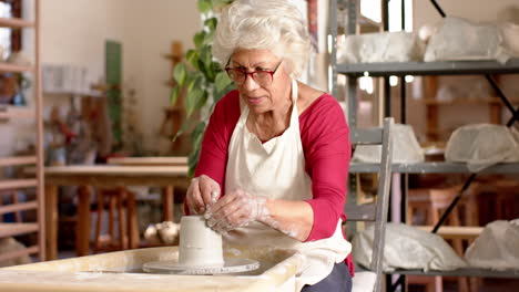 Senior-biracial-female-potter-with-gray-hair-using-potter's-wheel-in-pottery-studio,-slow-motion