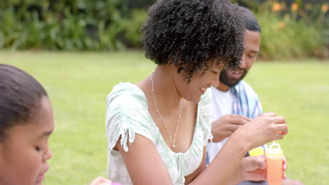 Glückliche-Afroamerikanische-Eltern-Und-Tochter-Pusten-Seifenblasen-Im-Garten,-Zeitlupe