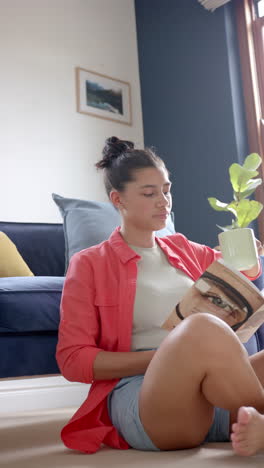 Vertical-video-of-happy-biracial-teenage-girl-sitting-on-floor-reading-book-with-tea,-slow-motion