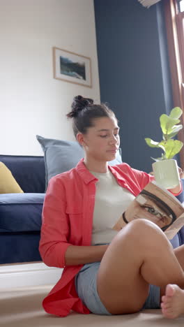 Video-Vertical-De-Una-Adolescente-Birracial-Feliz-Sentada-En-El-Suelo-Leyendo-Un-Libro-Con-Té,-Cámara-Lenta