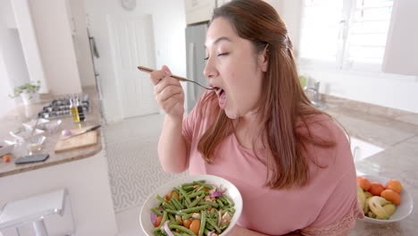 Happy-plus-size-biracial-woman-eating-vegetable-salad-standing-in-kitchen,-slow-motion